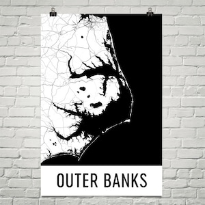 May include: A black and white map of the Outer Banks, North Carolina, displayed on a white poster. The map shows the coastline and roads in white against a black background. The words "OUTER BANKS" are printed in bold, black letters at the bottom.