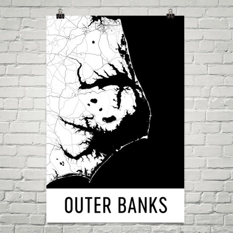 May include: A black and white map of the Outer Banks, North Carolina, displayed on a white poster. The map shows the coastline and roads in white against a black background. The words "OUTER BANKS" are printed in bold, black letters at the bottom.