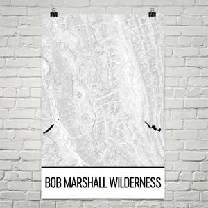 Puede incluir: Un póster de mapa topográfico en blanco y negro de Bob Marshall Wilderness. El mapa tiene una apariencia texturizada con líneas de contorno y detalles en negro. El póster está sujeto con clips contra una pared de ladrillos blancos.
