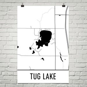 Op de afbeelding: Een zwart-wit kaartprint van Tug Lake. De kaart toont een gedetailleerde omtrek van het meer en de omgeving, met wegen en andere kenmerken. De woorden "TUG LAKE" zijn gedrukt in een vetgedrukte, schreefloze lettertype aan de onderkant van de print.