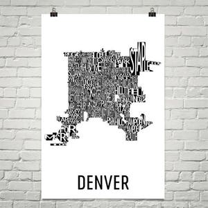 May include: A black and white poster featuring a map of Denver, Colorado, composed of neighborhood names. The word "DENVER" is printed in a bold, sans-serif font at the bottom of the poster. The poster is hanging on a white brick wall.