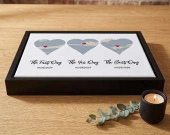 May include: A framed canvas print featuring three heart-shaped map designs in shades of blue, each with a red heart. Below the hearts are the phrases "The First Day", "The Yes Day", and "The Best Day" with dates. A lit candle and greenery are in the foreground.