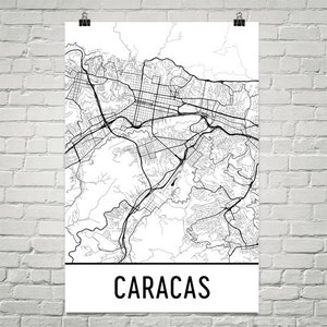 May include: A black and white line art map of Caracas, Venezuela. The map shows the city's streets and roads, as well as its surrounding mountains. The word "CARACAS" is printed in black at the bottom of the map.