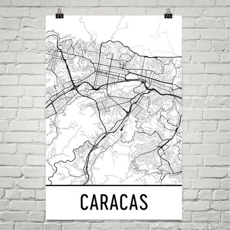 May include: A black and white line art map of Caracas, Venezuela. The map shows the city's streets and roads, as well as its surrounding mountains. The word "CARACAS" is printed in black at the bottom of the map.