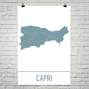 Puede incluir: Un póster con un mapa de Capri en color verde azulado apagado. El mapa detalla los contornos y carreteras de la isla. La palabra "CAPRI" está impresa en letras de bloque verde azulado en la parte inferior. El póster está sujeto con clips negros.