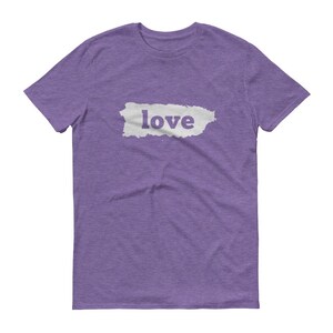 Puede incluir: Una camiseta morada con la palabra "love" impresa en blanco dentro de una forma rectangular blanca. La camiseta est&aacute; hecha de un material suave y tiene mangas cortas y cuello redondo.
