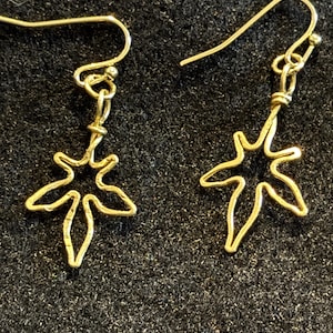 Könnte beinhalten: Ein Paar goldfarbene Drahtohrringe. Jeder Ohrring hat ein Ahornblatt-Design, das an einem Haken hängt. Die Ohrringe werden vor einem dunklen, strukturierten Hintergrund präsentiert.