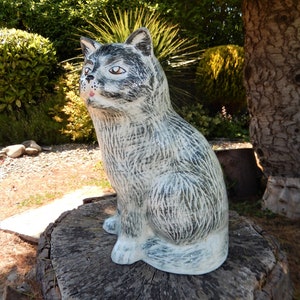 May include: A ceramic cat figurine with a white and grey painted finish. The cat is sitting on a tree stump in a garden setting.
