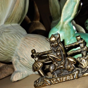 May include: A brass letter holder with an imp figure seated by a fence, the word "LETTERS" is on a banner. The background features ceramic rabbit figurines in green and white. The figurines add a whimsical touch.