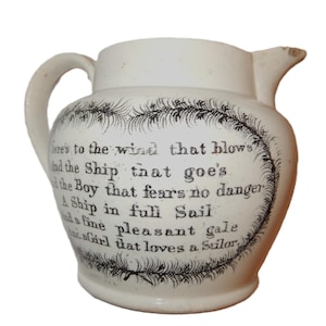 May include: Cream-coloured ceramic jug with a handle and spout. The jug has black text within a decorative border, including the phrases "Here's to the wind that blows" and "A Ship in full Sail."