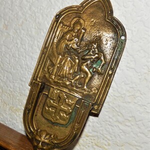 May include: A brass wall plaque featuring a relief sculpture of a woman in a robe holding a baby, with a small devil figure at her feet. The plaque has a decorative border and a coat of arms with a cross in the centre.