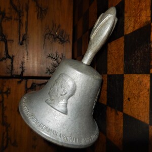 May include: A silver bell with a handle shaped like an aeroplane wing. The bell is inscribed with the text "Metal from German Aircraft" and has a profile of a man embossed on the side.