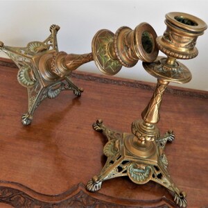 May include: Two antique brass candle holders with intricate detailing. Each holder has a square base with decorative feet and a central column with a candle cup. The brass has a warm, aged patina, and the design includes floral and geometric patterns.
