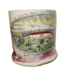 May include: A ceramic mug featuring a transfer print of a bridge over a river with sailing ships. The text reads View of the CAST IRON BRIDGE over the RIVER WEAR. Span 236 Feet. Height 100 Feet. Begun 1793. The base has a purple-toned glaze.