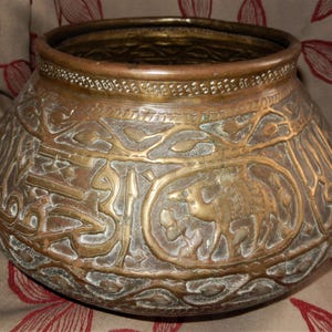 May include: A large, round, brass bowl with intricate embossed designs. The bowl features Arabic calligraphy and a stylized animal figure.