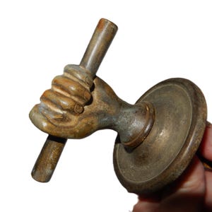 May include: Vintage bronze door handle, depicting a hand holding a rod. The handle has a circular base and a detailed hand design, with a weathered, aged appearance. The color is a mix of bronze and brown.