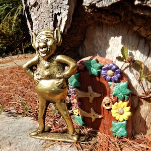May include: A gold-coloured gnome figurine with a wide smile stands next to a small, brown, wooden door with green ivy and colourful flowers. The door is decorated with a keyhole and two metal handles.
