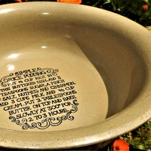 May include: A beige ceramic bowl with a recipe for rice pudding printed in black. The recipe includes ingredients like sugar, salt, nutmeg, cinnamon, milk, and cream. The bowl is surrounded by orange flowers.