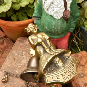 May include: A brass door knocker featuring a pixie figure holding a mushroom. The knocker is attached to a bell with a chain. The knocker is engraved with the words "Exmoor Pixie".