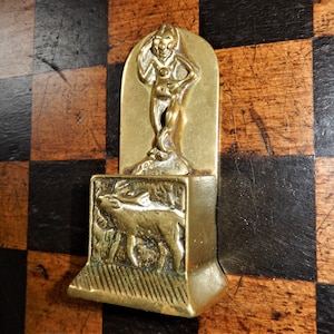 May include: A brass wall mounted match holder with a small pig design and a figure of a woman holding a flower. The holder has a textured surface and is mounted on a wooden surface.