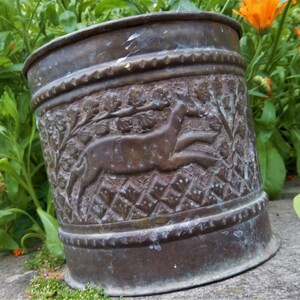 May include: A vintage copper planter featuring an embossed design of a deer running through a woodland scene. The planter has a dark brown patina and is in good condition.