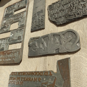 May include: A selection of antique metal printing blocks, featuring diverse designs and text. The blocks vary in shape and size, showcasing intricate relief patterns. The background is a light wood grain.