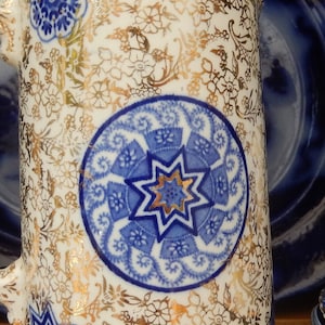 May include: A white ceramic jug with a blue and gold floral pattern. The jug has a lid and a handle. The pattern includes a large blue and gold star in the centre.