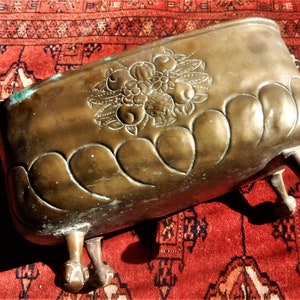 May include: A vintage brass planter with a floral design and three feet. The planter is on a red rug.