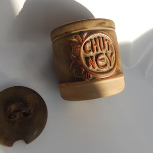 May include: A brown ceramic jar with a lid, decorated with a brown and orange design. The jar says "CHUTNEY" in orange letters.