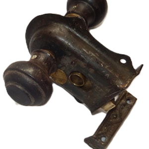 May include: A dark brown wooden door knob set with a black metal mortise lock mechanism. The hardware includes a rectangular metal strike plate with a geometric embossed pattern and a circular brass keyhole insert on the main lock body.