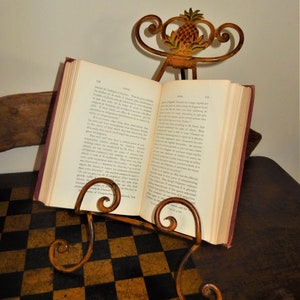 May include: A gold-toned metal book stand with a pineapple design on the top. The stand is holding open a book with text in French. The stand is sitting on a brown and black checkered surface.