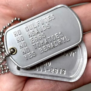 K&ouml;nnte beinhalten: Zwei silberne Hundemarken an einer silbernen Kette. Die obere Marke lautet "Keine Sulfite, Keine NSAR, Keine Eier, Keine Tomaten, Ja Benadryl". Die untere Marke lautet "JJJ-2813".