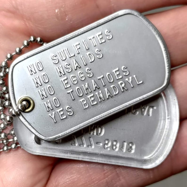 K&ouml;nnte beinhalten: Zwei silberne Hundemarken an einer silbernen Kette. Die obere Marke lautet "Keine Sulfite, Keine NSAR, Keine Eier, Keine Tomaten, Ja Benadryl". Die untere Marke lautet "JJJ-2813".