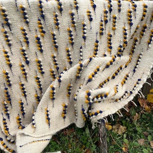 Puede incluir: Una manta de tejido blanco con un patrón geométrico de pompones amarillos y azules. La manta está drapeada sobre una rama con hojas de otoño en el fondo.