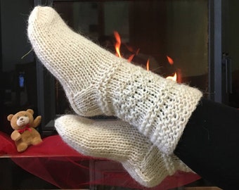 May include: A pair of white knitted socks with a cable knit pattern, worn by a person sitting in front of a fireplace.