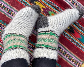 May include: A pair of white wool socks with green and beige stripes. The socks are being worn by a person sitting on a colourful rug.