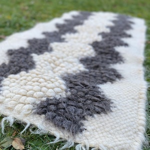 Puede incluir: Una alfombra de lana blanca y gris con un patrón geométrico. La alfombra está tejida a mano y tiene una textura esponjosa.