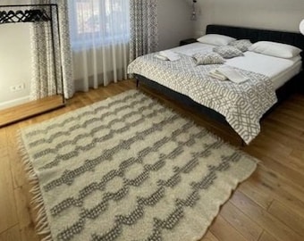 May include: A bedroom with a large bed with a white and gray patterned comforter. The bed has a black headboard and is in front of a window with white curtains. There is a white rug on the floor and a wooden ceiling with exposed beams.