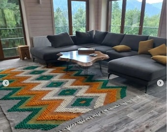 May include: A gray sectional sofa with yellow throw pillows sits on a wood floor with a large, colorful rug. The rug has a geometric pattern of orange, green, and white. A wooden coffee table with a live edge sits in front of the sofa.