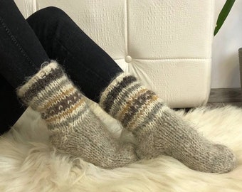 May include: A pair of gray wool socks with brown and beige stripes. The socks are worn by a person sitting on a white fluffy rug.
