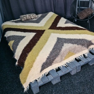 Puede incluir: Una manta tejida con un patrón geométrico en tonos marrones, blancos y verdes. La manta está drapeada sobre un marco de cama de palets de madera.