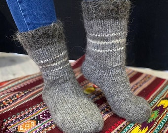 May include: A pair of grey wool socks with white stripes. The socks are knitted and have a fluffy texture. The socks are on a colourful rug.