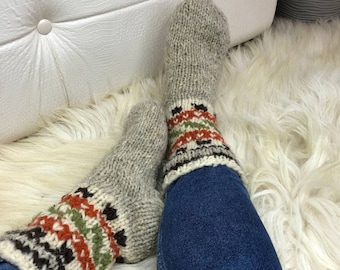 Könnte beinhalten: Ein Paar graue Wollsocken mit bunten geometrischen Mustern an den Bündchen. Die Socken werden an den Füßen einer Person getragen und liegen auf einer weißen, flauschigen Oberfläche.