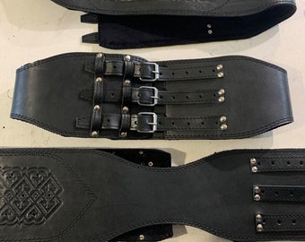 Puede incluir: Tres cinturones de cuero negro con diferentes diseños. El cinturón superior tiene un patrón de nudo celta y está unido a un respaldo de tela negra. El cinturón del medio tiene tres filas de hebillas plateadas y está decorado con tachuelas plateadas. El cinturón inferior tiene un patrón de nudo celta y está decorado con tachuelas plateadas.