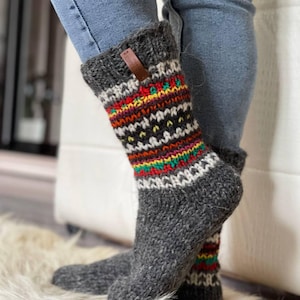 May include: A pair of gray knit socks with a colorful geometric pattern. The socks feature bands of red, yellow, green, and white. A small brown tag is attached to the cuff. The socks are worn with blue jeans.