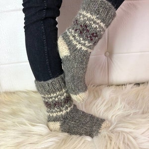 May include: Grey knitted socks with a white and burgundy geometric pattern. The socks have white heels and toes. These thick socks appear warm, ideal for colder weather. The socks are made of a thick yarn.