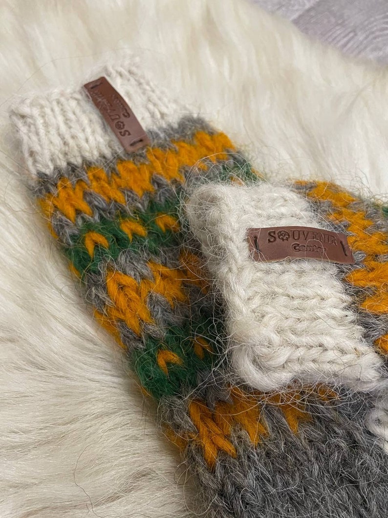 May include: A pair of knitted fingerless gloves with a white, grey, green, and yellow striped pattern. The gloves have a brown leather tag with the text "SOUVENIR" on them.