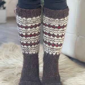 May include: A pair of knee-high knitted socks. The socks are primarily brown with a patterned design in white and red. The socks are worn over black pants and rest on a white, fluffy surface.