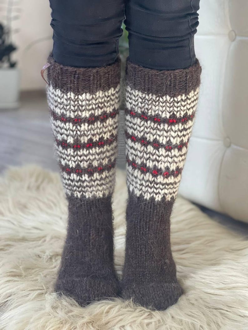 May include: A pair of knee-high knitted socks. The socks are primarily brown with a patterned design in white and red. The socks are worn over black pants and rest on a white, fluffy surface.