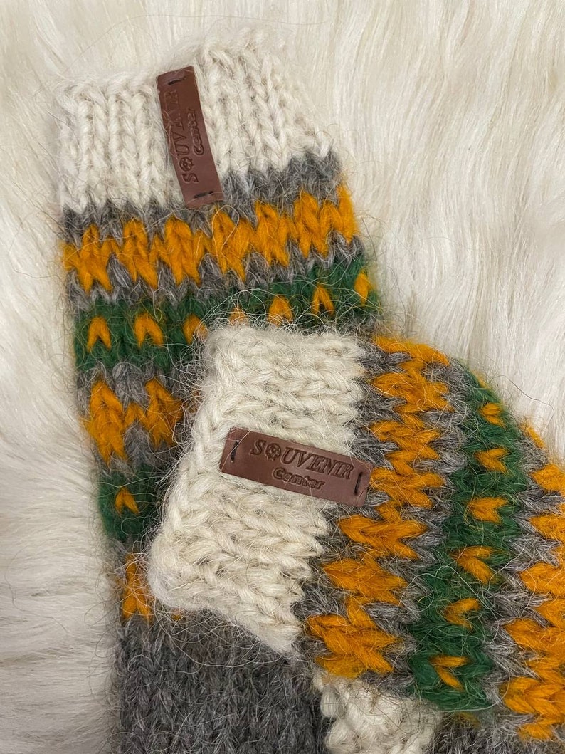 May include: A pair of knitted wool socks with a colourful geometric pattern in shades of white, grey, green, and orange. The socks have leather tags that say "Souvenir" and "Canada".